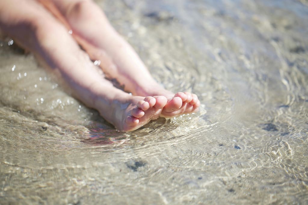 hidroterapia-sensorial-temperatura-ambiente-bio-yin Primer plano de pies descalzos en la orilla del agua cristalina, representando la hidroterapia sensorial y la reconexión natural.