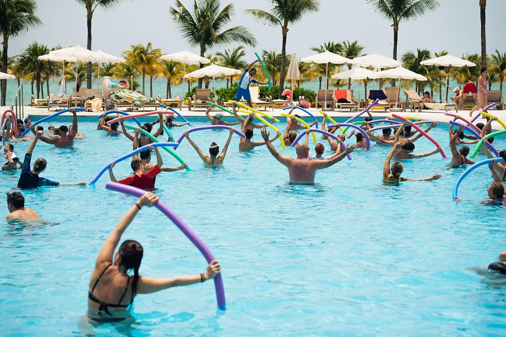 Beneficios-ejercicio-acuatico Grupo de personas realizando ejercicios de hidroterapia y zumba acuática en una piscina para fortalecer músculos y tendones sin impacto.