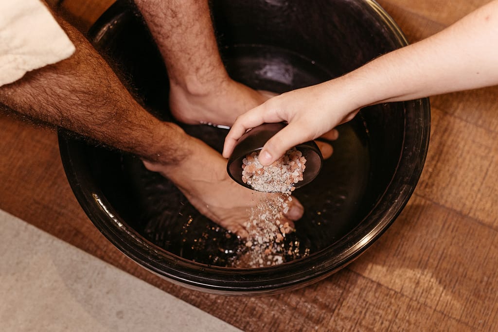 beneficios-sales-epsom-baño-pies Una mano virtiendo sales de baño rosadas o sal de Epsom en un cuenco con agua tibia y pies sumergidos para un pediluvio relajante.