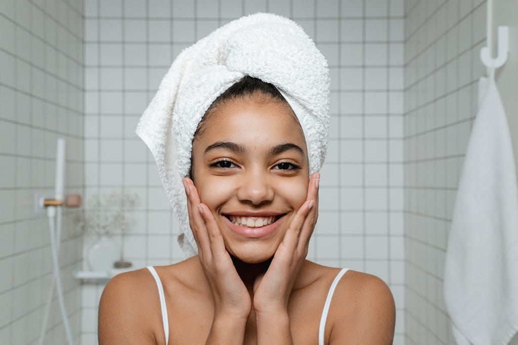 Gimnasia vascular y baños de contraste para el cutis Joven sonriente con una toalla en el cabello sujetando su rostro en el baño tras una rutina de limpieza facial e hidroterapia.