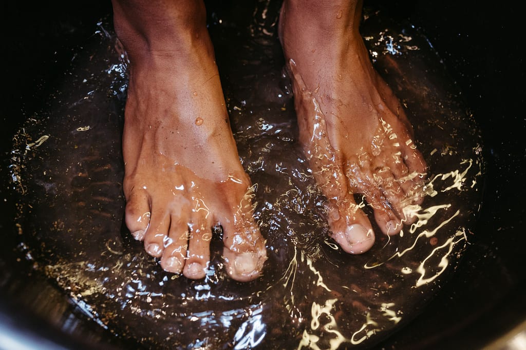guia-practica-pediluvio-en-casa Pies sumergidos en agua tibia dentro de un recipiente oscuro para realizar un pediluvio o baño de pies terapéutico.