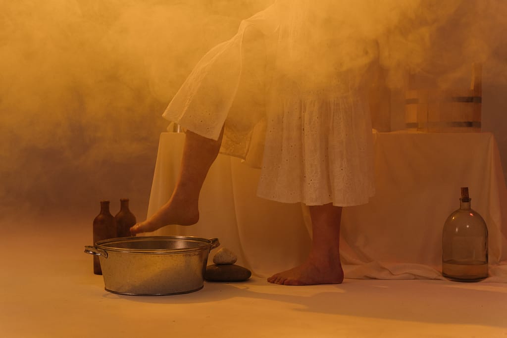 pediluvio-terapias-naturales-bienestar Persona vestida de blanco preparando un baño de pies en un balde metálico en una habitación llena de vapor cálido y botellas de vidrio terapéuticas.