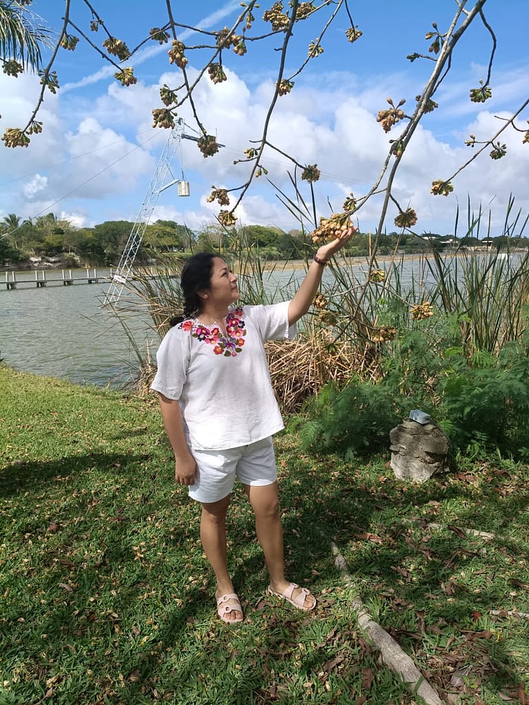 Posando-Lago-Yucatán Mujer joven posando junto a un lago, tocando las ramas de un árbol con frutos, vistiendo una blusa blanca bordada.